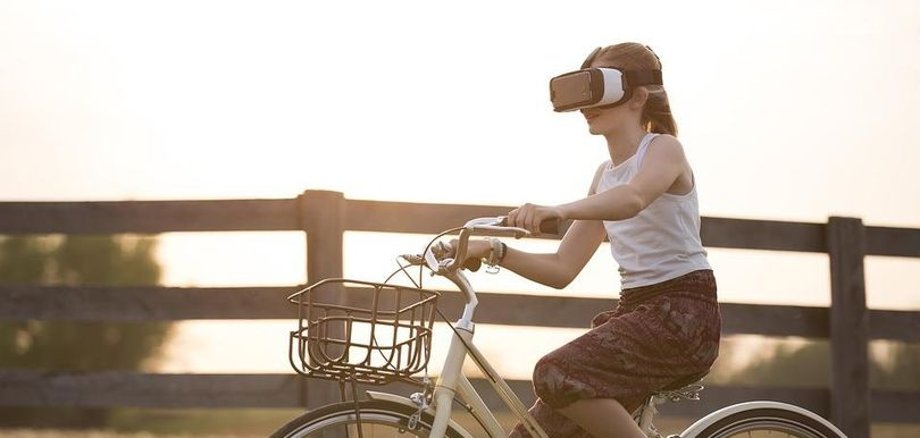 Mädchen hat VR Brille auf und fährt Fahrrad vor einem ländlichen Hintergrund (Weidenzaun)