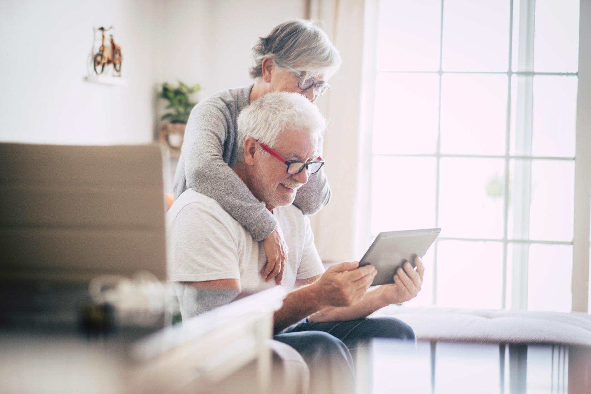 couple of seniors smiling and looking at the same tablet hugged on the sofa - indoor, at home concept - caucasians mature and retired man and woman using technology